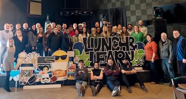 Members of Lunch & Learn Networking gathered for a group photo at a Wednesday meeting in Hudson, Wisconsin, standing and seated around a large hand-lettered Lunch & Learn sign.